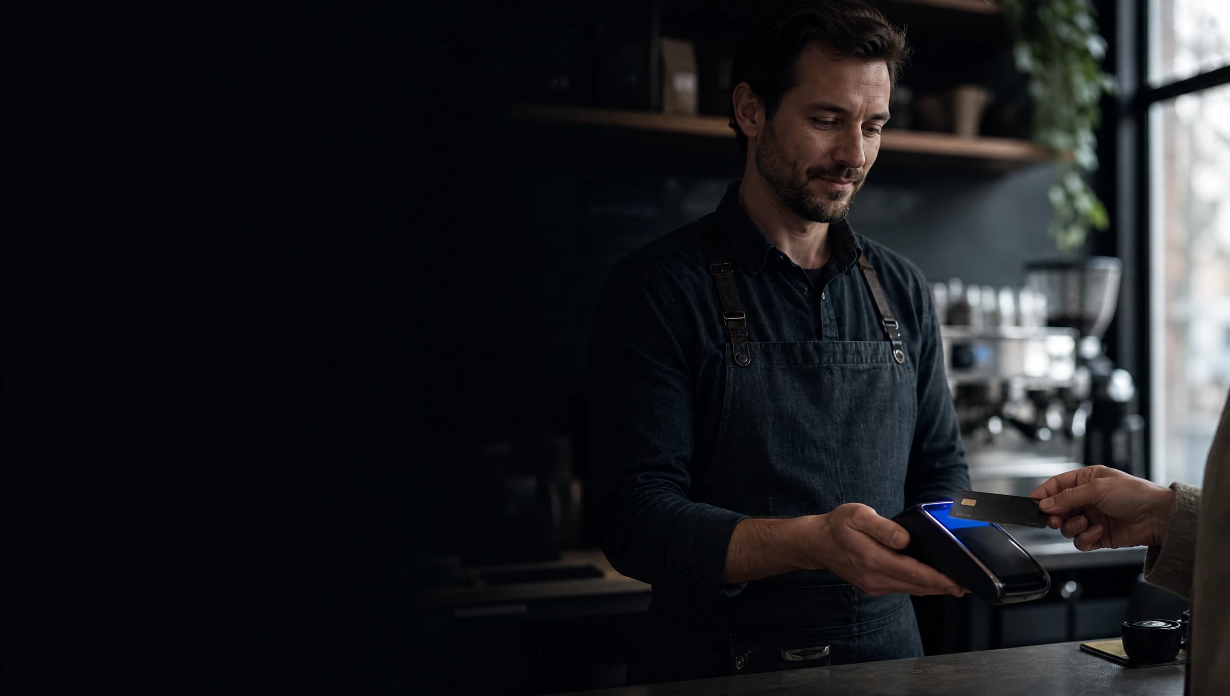 Café owner in an apron holding out a payment terminal as a customer taps a bank card at the counter